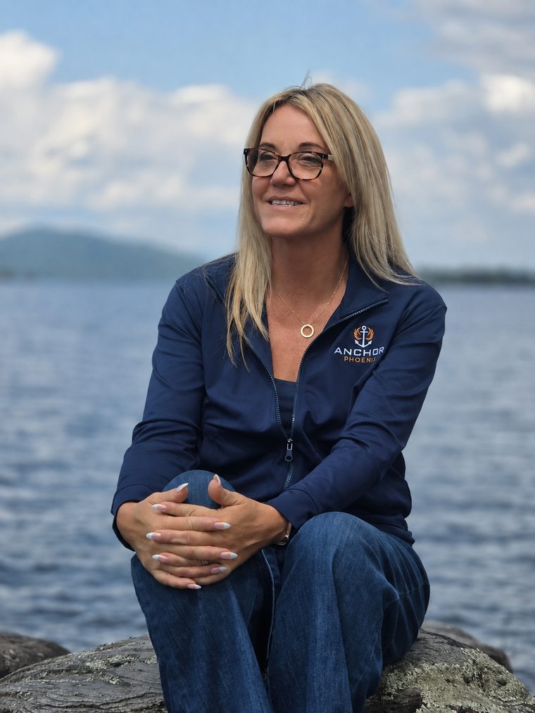 Beth LaFontaine, founder of AnchorPhoenix, seated on a rock beside a lake.